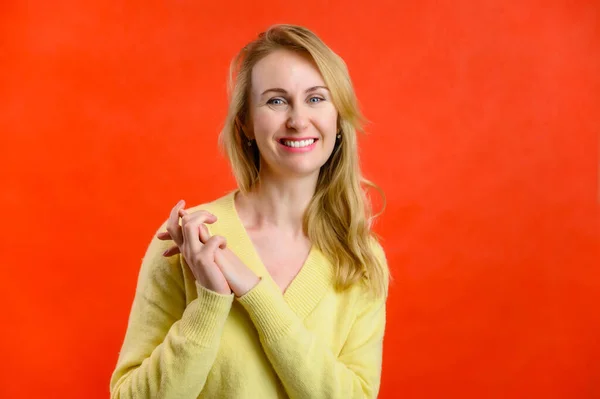 Mulher loira de 40 anos de idade em um suéter amarelo mostra alegria com as mãos em um fundo vermelho — Fotografia de Stock