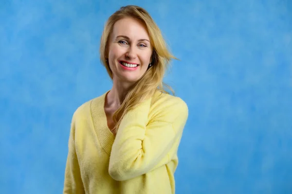 Bonita mujer rubia de 40 años en un suéter amarillo muestra un estilo de vida alegre sobre un fondo azul — Foto de Stock