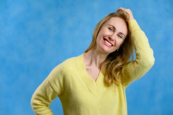 Mulher loira bonita 40 anos em uma camisola amarela mostra a alegria da vida em um fundo azul — Fotografia de Stock