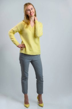 Sarı kazaklı, beyaz pantolonlu, gülümseyen sarışın bir kadının tam boy portresi.