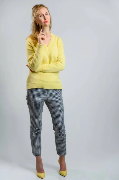 Фото на белом фоне в полный рост улыбающейся белокурой белокурой женщины в желтом свитере и серых брюках, думающей с ручкой в руках — стоковое фото