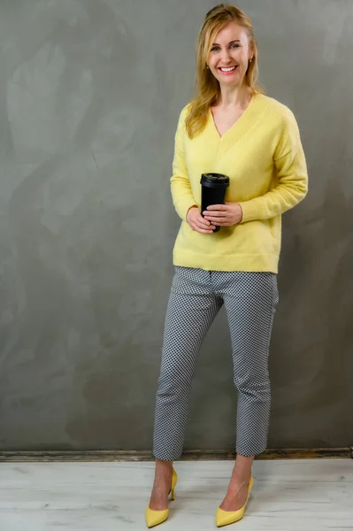Blond blank vrouw in een gele trui en grijze broek staat recht met koffie in haar handen op een grijze achtergrond in volle groei gelukkig glimlachen — Stockfoto