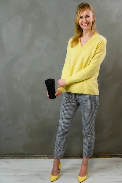 Кавказька блондинка з посмішкою в жовтому светрі і сірих штанях стоїть прямо з кавою в руках на сірому фоні в повній довжини. — стокове фото