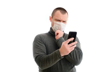 Model tam kameranın önünde duruyor. Akıllı telefon ile duygular ve el konsepti ile beyaz arka planda gri kazaklı, tıbbi maskeli beyaz adamı izole edin.