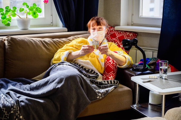 Sick woman in a bathrobe with a medical mask lies in the home interior on the couch. She takes medication pills.