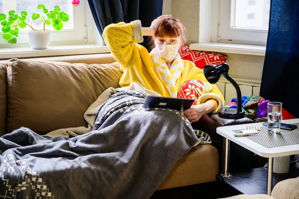 Photo of an adult woman with a viral infection, lying at home on a sofa in a medical mask. Uses a tablet computer