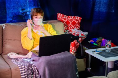 Evin içinde karantina altında maske takan bir kadının fotoğrafı. Model çalışıyor, internet üzerinden dizüstü bilgisayar kullanıyor..