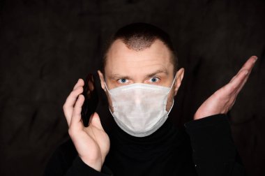 Siyah arka planda elinde telefonla tıbbi maske takan yetişkin bir adamın yakın plan portresi.