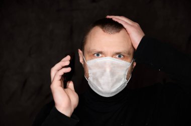 Siyah arka planda elinde telefonla tıbbi maske takan yetişkin bir adamın yakın plan portresi.