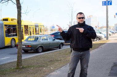 Siyah ceketli, güneşli bir günde şehirde gözlüklü yakışıklı bir adamın sokak fotoğrafı parmaklarını kenarda gösteriyor.