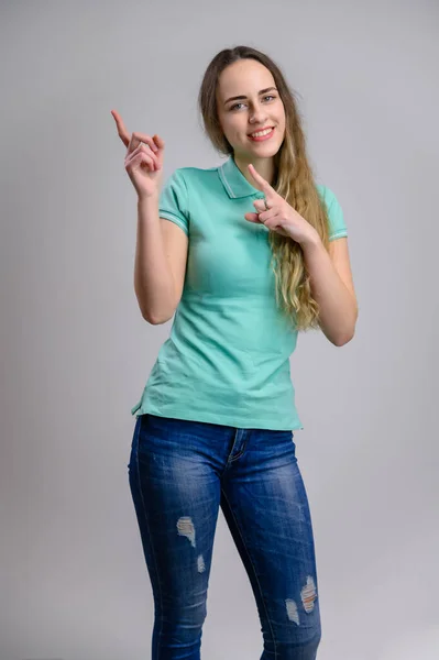 白い背景にターコイズブルーのジーンズのブラウスに長い髪を持つかなり若い女性の垂直写真 モデルは指を側面に向け — ストック写真