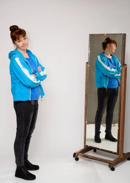 Vertical photo of a woman in a blue blouse and jeans standing in front of a mirror on a white background