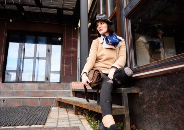 Fotoğraf portresi, şehirde bej ceketli esmer bir kız güneşli bir havada bir dükkan penceresinin arka planında oturuyor.