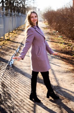 Çok güzel gülümseyen sarışın beyaz bir kızın portresi. Uzun, güzel saçları var. Parkta pembe bir palto içinde güneşli bir günde bir ağaca yaslanmış..