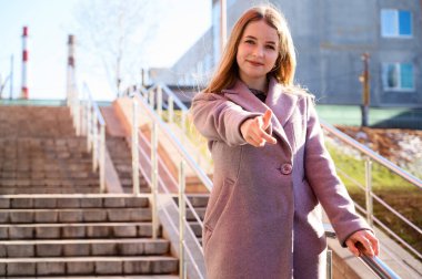 Güneşli bir günde, binanın arka planında pembe bir palto giyen, uzun, güzel saçlı, sarışın, gülümseyen, beyaz bir kızın fotoğrafı. Model poz vermekten mutlu..