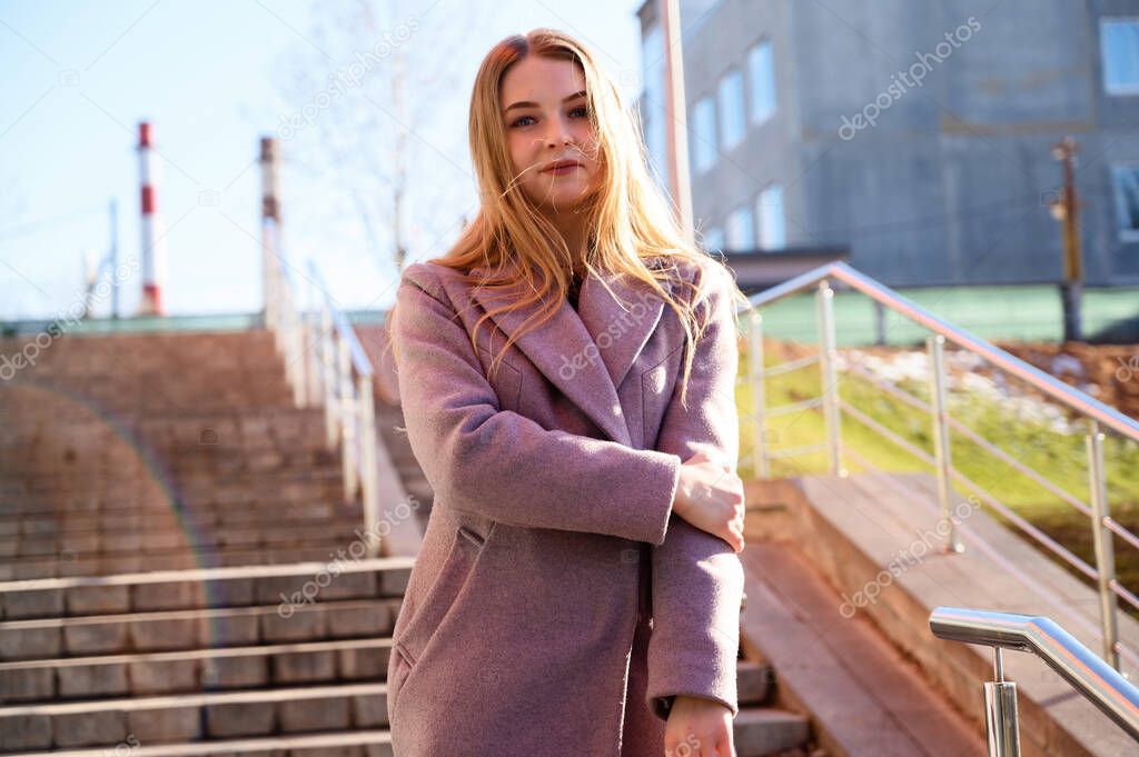 A pretty model is happy to pose. Photo of a happy smiling Caucasian blonde girl with long beautiful hair outdoors in a pink coat on the background of the steps of the building in sunny daytime.
