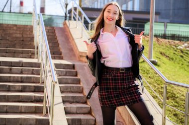 Şehrin arka planında, güneşin altında duyguları olan mutlu, gülümseyen güzel bir kızın bahar dikey fotoğrafı. Kamera önünde poz veren güzel bir model..