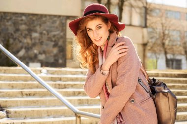 Şık, alımlı, çekici, uzun saçlı güzel bir kadının fotoğrafı. Güneşli bir bahar şehrinde. Binanın arka planında bir model duruyor.