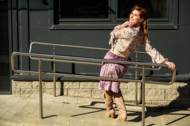 Güzel, modaya uygun, uzun saçlı bir kadının tam boy fotoğrafı. Baharda, güneşli bir havada, bir binanın arka planında..