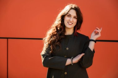 Oldukça şık, mutlu esmer bir kadının fotoğrafı. Uzun, güzel saçlı, şehirde takım elbiseli, turuncu arka planda, güneşli bir havada bina duvarında.