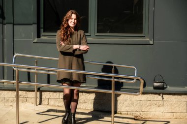 Şık, mutlu, uzun, güzel saçlı bir esmer kadının fotoğrafı. Şehrin arka planında, güneşli bir havada binanın merdivenlerinde, takım elbiseli bir kadın.