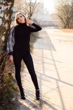 Uzun saçlı, kentte gülümseyen ve pencereli bir iş binasının duvarına dayanan zarif bir genç kadının tam boy portresi. Baharda güneşli ve açık havada çekilmiş bir fotoğraf..