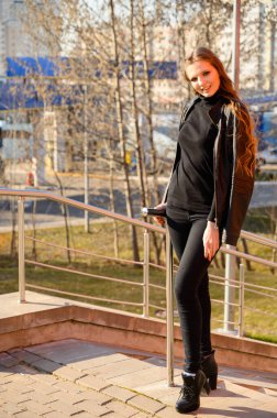 Şehrin arka planında uzun saçlı ve gülümseyen zarif bir genç kadının tam boy portresi. Baharda güneşli ve açık havada çekilmiş bir fotoğraf..