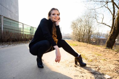 Şehirdeki bir yolun arka planında uzun saçlı ve gülümseyen güzel bir genç kadının portresi. Baharda güneşli ve açık havada çekilmiş bir fotoğraf..