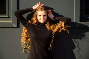 Pencereli bir iş binasının arka planında gülümseyen uzun saçlı güzel bir genç kızın şehir portresi. Baharda güneşli ve açık havada çekilmiş bir fotoğraf..