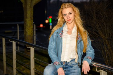 Sarışın bir kızın portresi. Gece şehirde, gün batımında, arka planda ışıkların üzerinde.