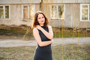 Şık, zarif, esmer bir kızın, şehrin bahar güneşinde arka planında pencerelerin önünde poz vermesi.