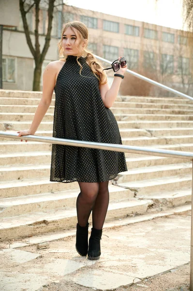 今年春天 城市一座室外建筑的台阶上站着一位穿着黑色衣服的金发美女的画像 — 图库照片
