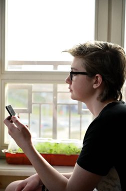 Gözlüklü beyaz adam, pencerenin karşısında oturan akıllı telefonu kullanıyor.