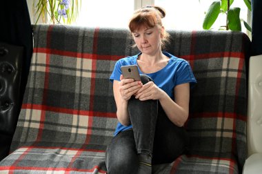 Karantinada mavi tişörtlü yetişkin bir kadının akıllı telefonuyla konuşurken bir kanepede otururken çekilmiş fotoğrafı.