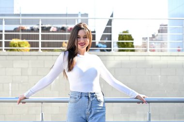 Beyaz bluzlu ve mavi kotlu güzel bir esmer kızın beyaz portresi, güneşli bir bahar gününde, bir binanın gri duvarının arkasında tebessümle korkulukların yanında duruyor..