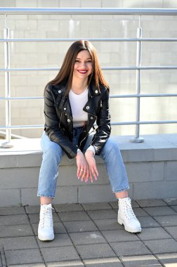 Siyah ceketli, mavi kotlu, güneşli bir bahar gününde korkulukta oturan güzel esmer bir kızın dikey beyaz portresi..