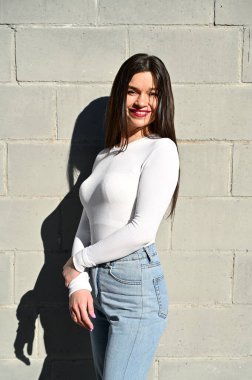 Beyaz bluz ve mavi kot giyen güzel esmer bir kızın dikey beyaz portresi şehrin bahar günü bir binanın duvarında duruyor. Model poz vermekten mutlu..