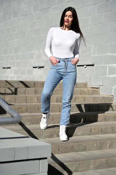 白いブラウスと青いジーンズを着た白人のブルネットの女の子の写真は 晴れた春の日の建物の灰色の壁の背景に笑顔で階段に立っています — ストック写真