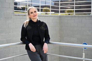 Öğleden sonra açık havada, arka planda merdivenlerin korkuluğunda güzel, beyaz bir sarışının fotoğrafı. Pantolon ve ceket giyen modaya uygun bir manken gülümseyerek poz verir.