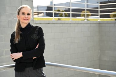 Pantolon ve ceketli, gülümseyerek poz veren modaya uygun bir model. Açık havada güneşli bir günde, arka planda, merdivenlerin korkuluğunda güzel sarışın beyaz bir kadının fotoğrafı..