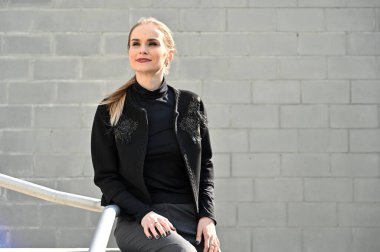 Açık havada güneşli bir günde, arka planda merdivenlerin korkuluğunda güzel, beyaz, sarışın bir kadının fotoğrafı. Pantolon ve ceket giymiş bir manken gülümseyerek poz verir..