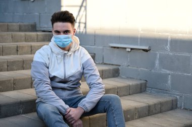 Açık havada, güneşli bir günde gri bir arka planda tıbbi maske takan beyaz bir gencin portresi.