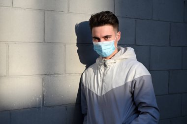 Açık havada, güneşli bir günde gri bir arka planda tıbbi maske takan beyaz bir gencin portresi.