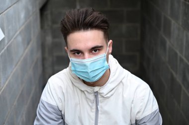 Açık havada, şehirdeki bir binanın bodrumunun arka planında açık renk ceketli esmer bir adamın portresi. Koruyucu tıbbi maskeli bir model..