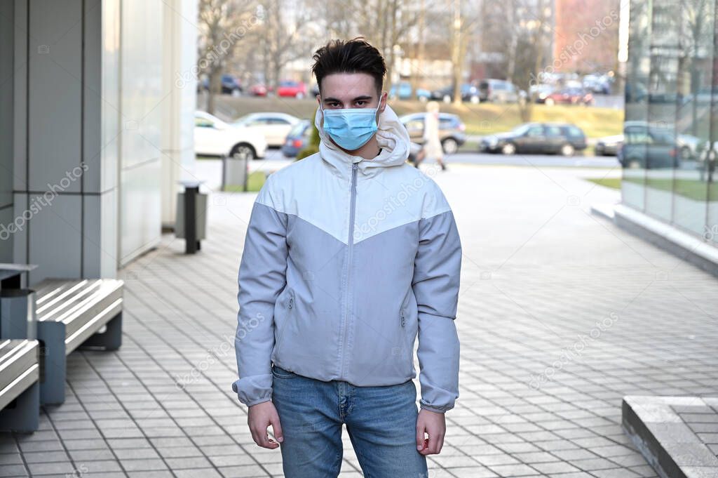 Photo Caucasian young man in a medical mask in a light jacket outdoors on a city background on a sunny day