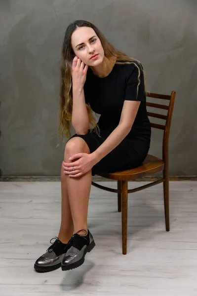一个身穿黑色长发长裙的年轻漂亮女子的垂直肖像 模特坐在灰色的背景上 坐在一把时髦的椅子上 — 图库照片
