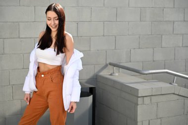 Fotoğraf güneşli bir bahar gününde çekildi. Açık havada, tıbbi maskeli, bej pantolonlu, beyaz ceketli, esmer bir kızın portresi şehrin gri duvarında..