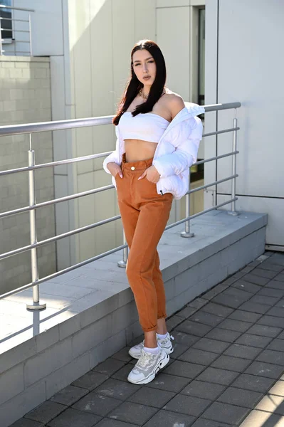 Vertical Full length photo outdoors of a young brunette girl in beige trousers and a white jacket on the background of the railing of the building s stairs in the city. Made on a sunny spring day.