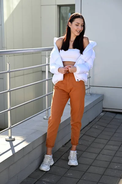 Vertical Full length photo outdoors of a young brunette girl in beige trousers and a white jacket on the background of the railing of the building s stairs in the city. Made on a sunny spring day.