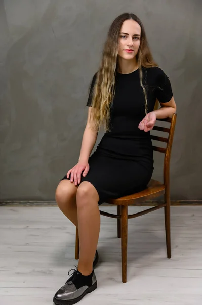 工作室里一个穿着黑色裙子 灰色背景的年轻漂亮女人的垂直照片 一个长头发的模特坐在椅子上 — 图库照片